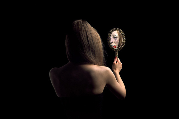 Young woman looking into a face mirror with her back to the camera, her reflection is the mask of an old woman